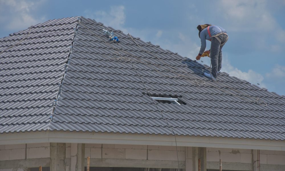 A construction worker wearing a full body safety belt is installing tile on the roof of a large house. By using corrugated ceramic tiles on a strong steel roof frame.